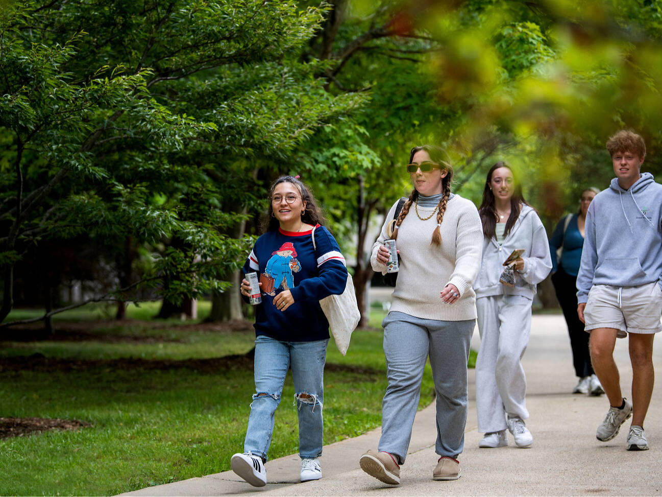 A group of young people stroll along a tree-lined path, casually dressed and holding drinks.